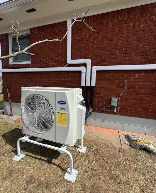 HVAC heat pump unit mounted on ground outside red brick building with white PVC piping and electrical conduit