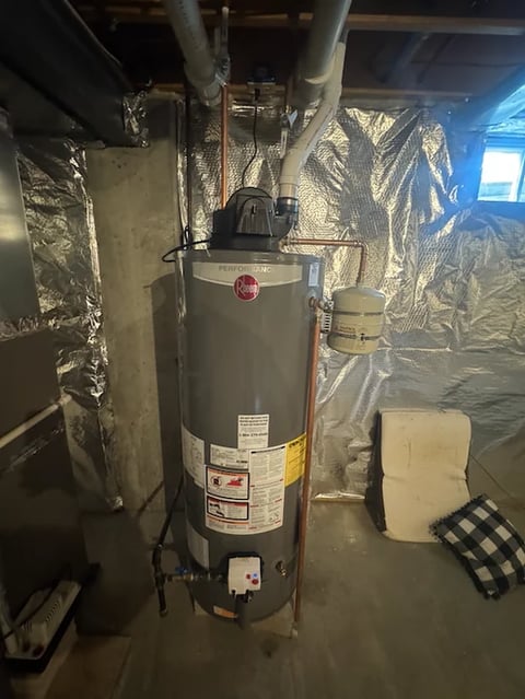 Gas water heater in basement with insulation wrap and pipes, surrounded by storage boxes and foam padding