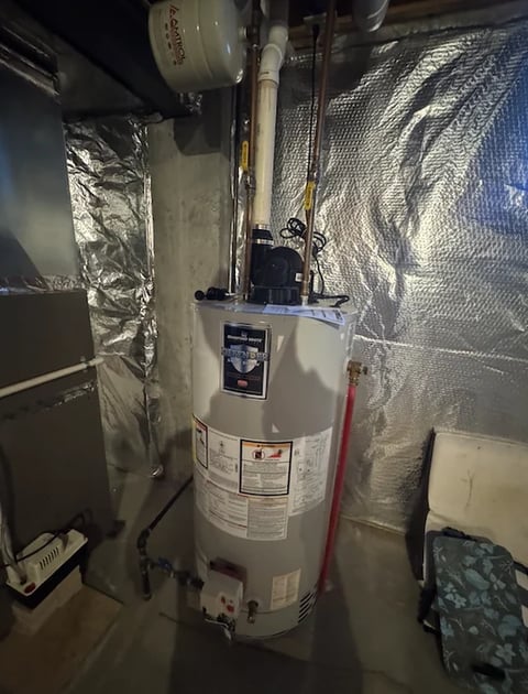 White cylindrical water heater in basement with insulation wrap, pipes, and red valve visible